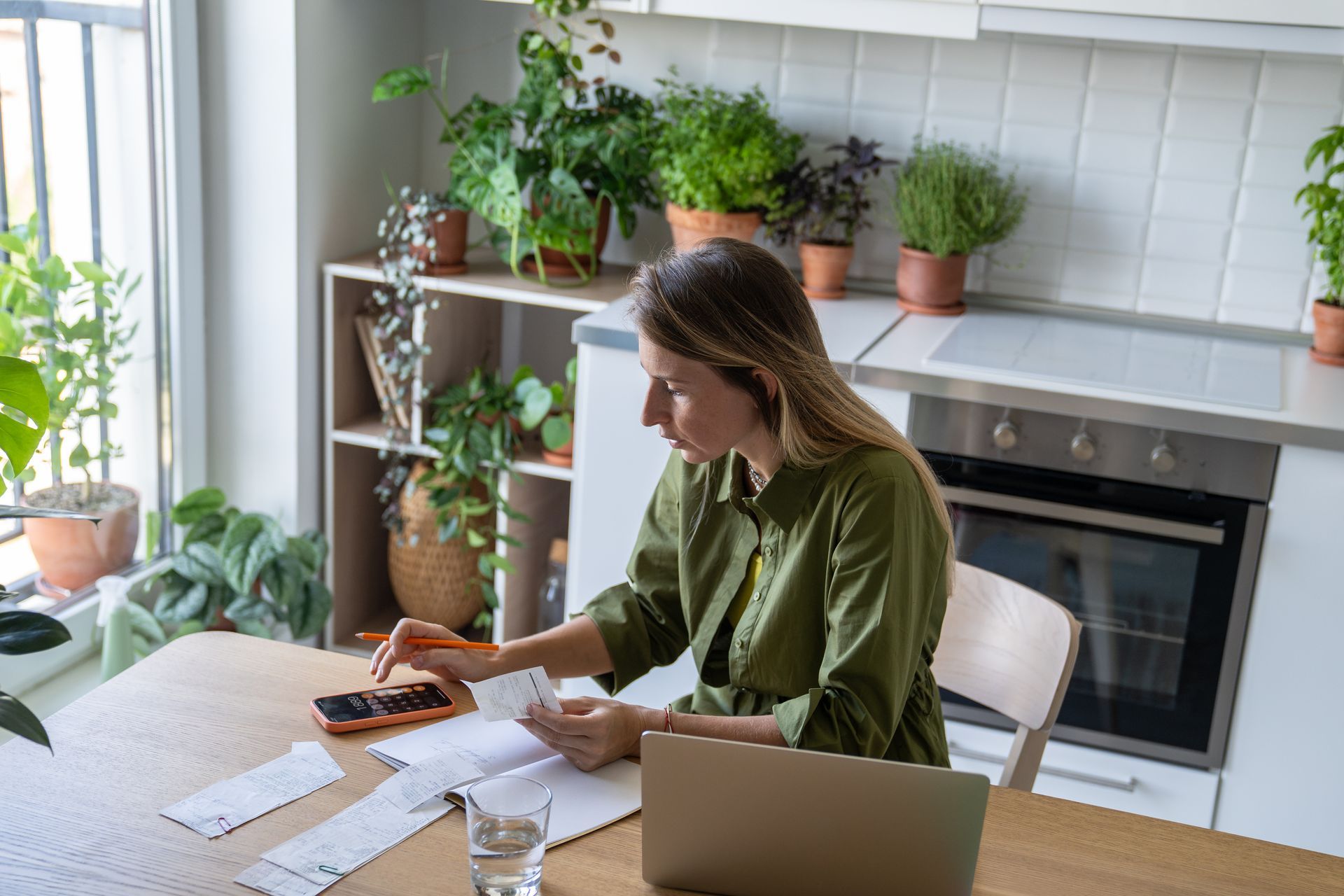 Une femme calcule des factures à une table, entourée de plantes et d'un ordinateur portable dans une cuisine.