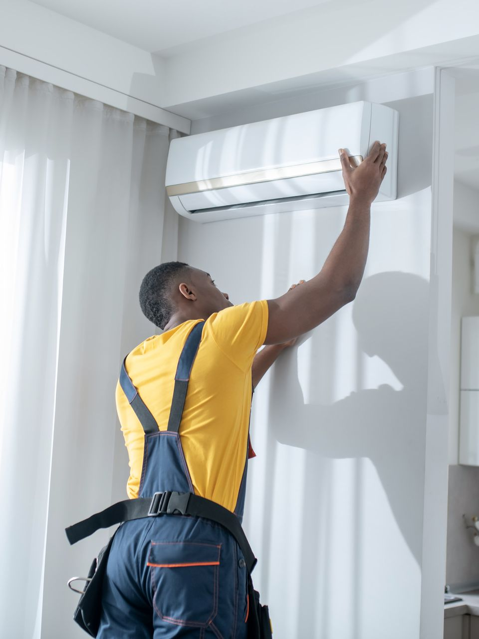Un homme en salopette installe un climatiseur sur un mur blanc.