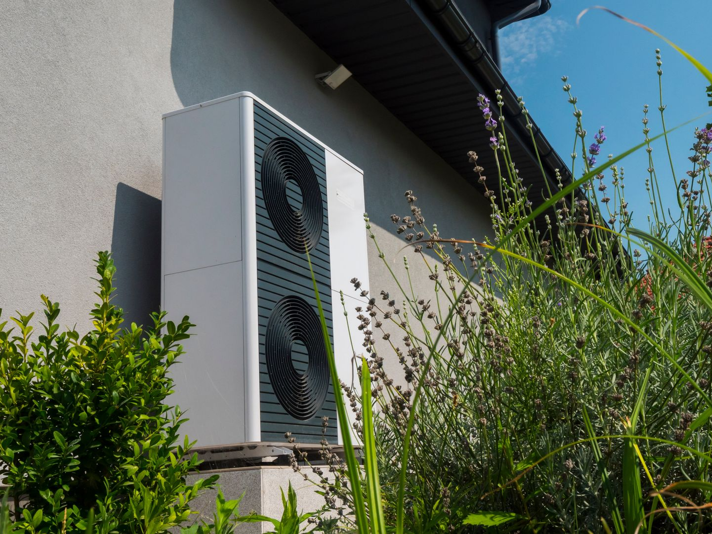Unité de pompe à chaleur blanche fixée sur le côté d'une maison, à côté de buissons.