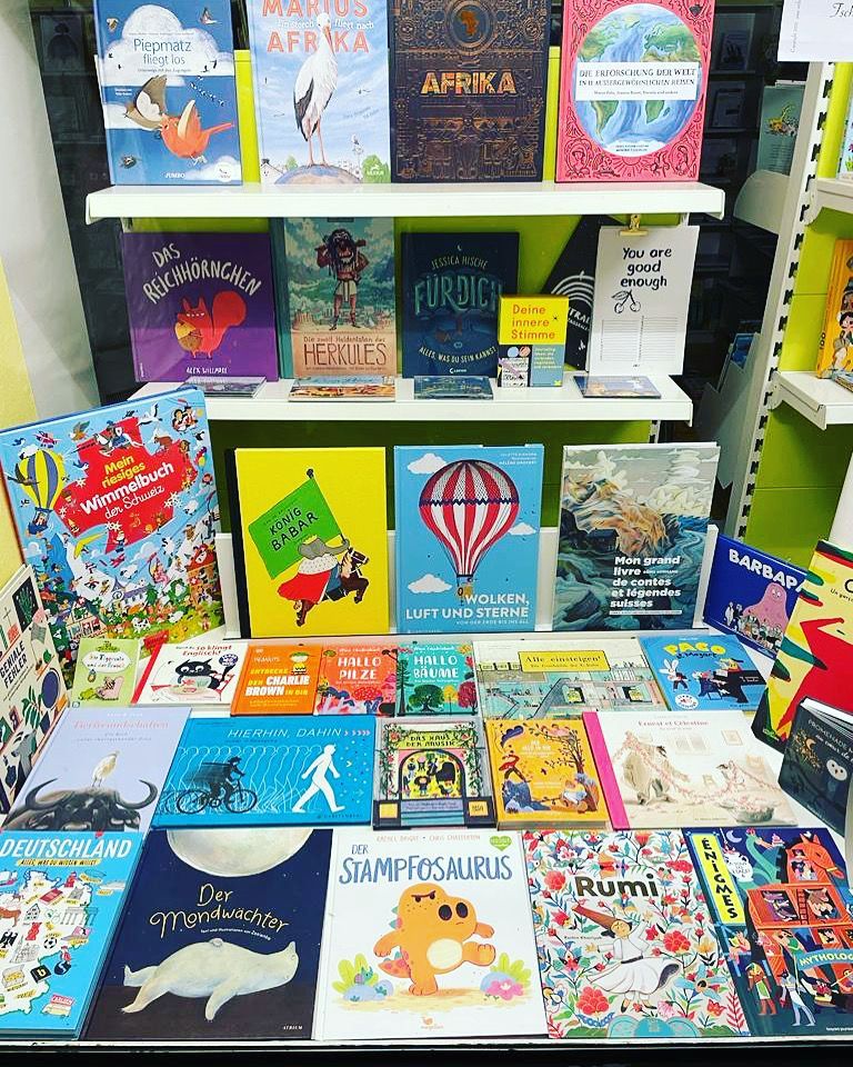 Mädchen stöbert in einem hell erleuchteten Buchladen in Büchern. Sie trägt ein gemustertes Hemd und Shorts sowie hellblaue Sandalen.