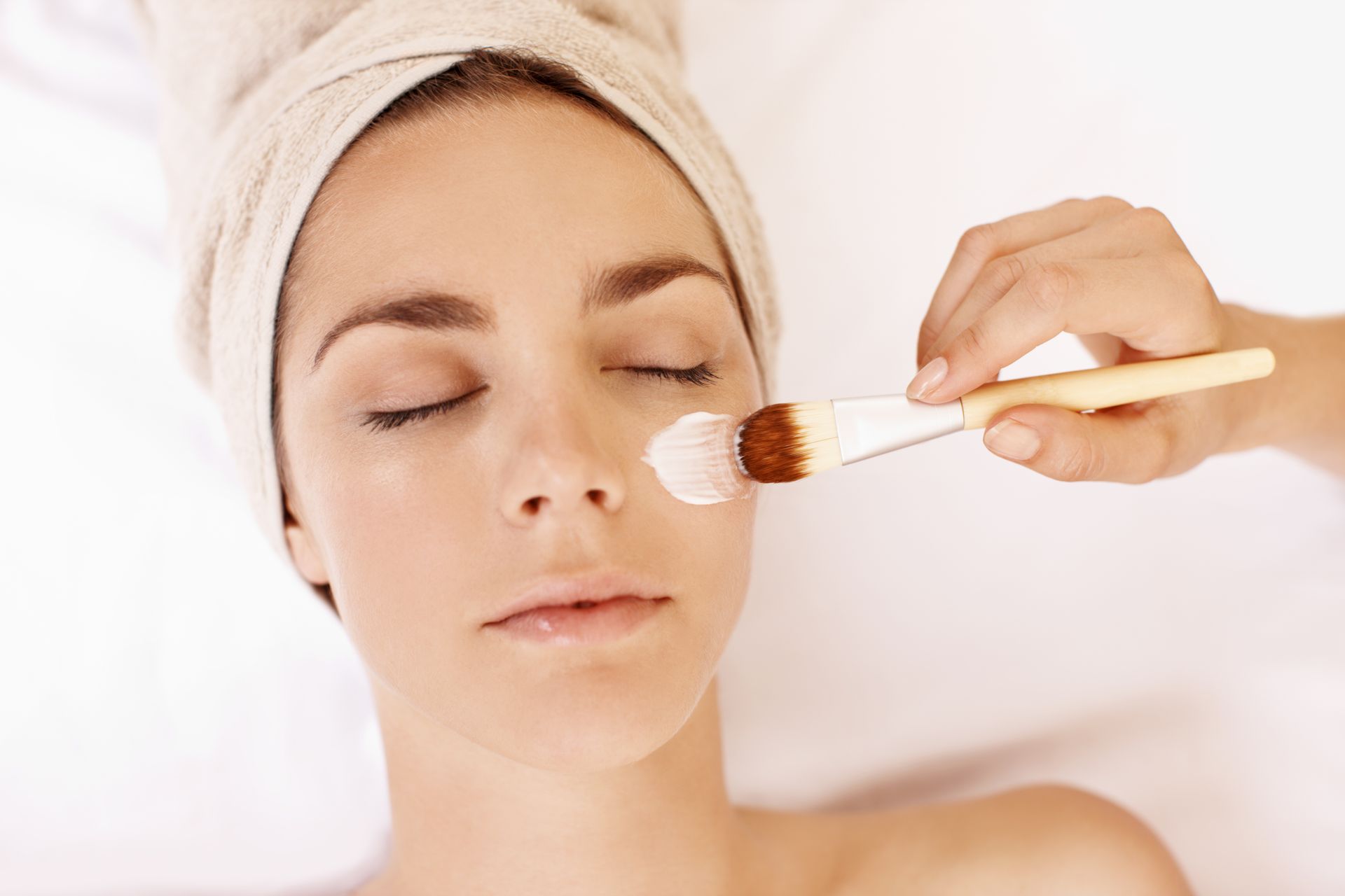Femme avec une serviette sur la tête recevant un soin du visage avec un pinceau sur la joue