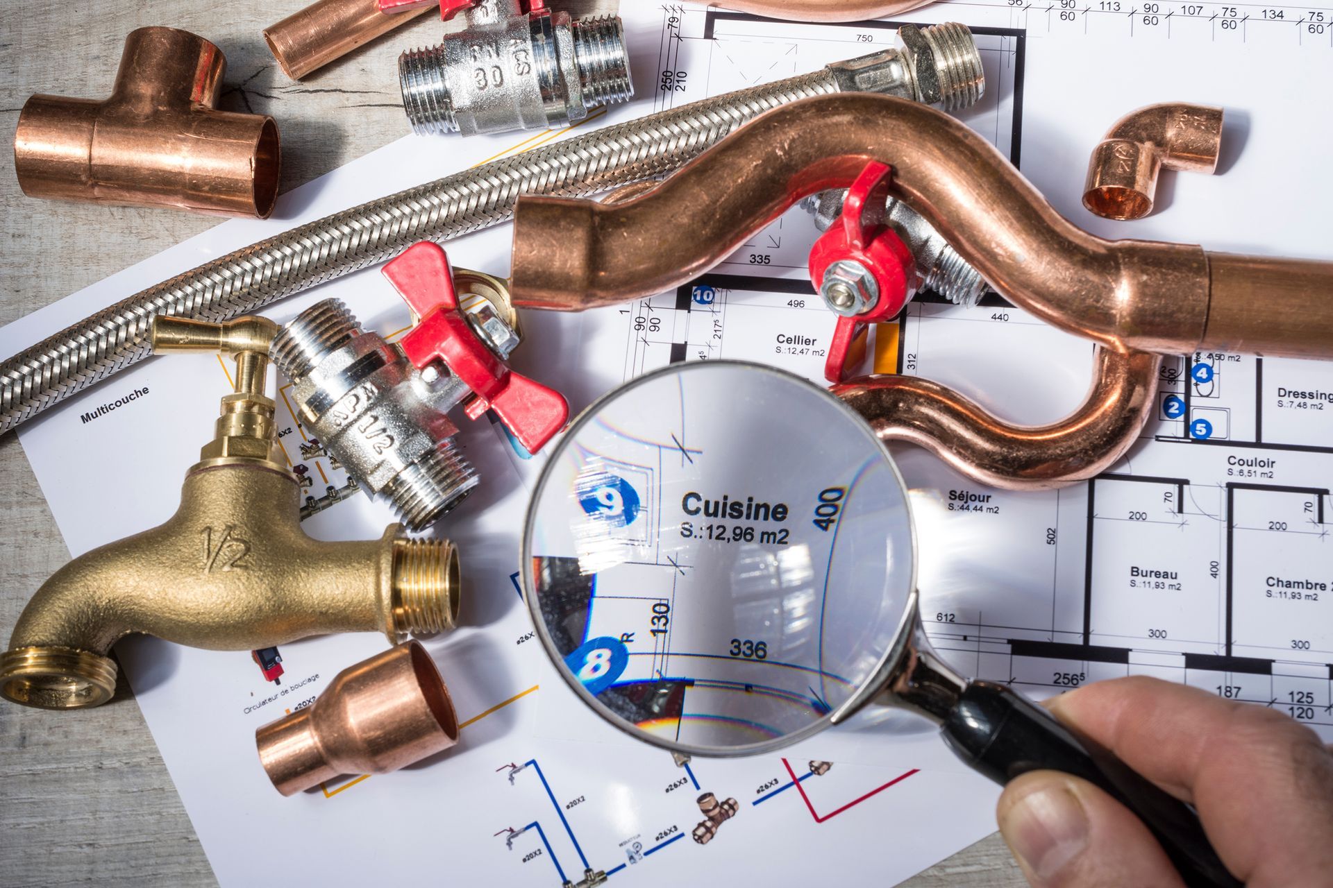 Une loupe examine le plan d'une cuisine entourée de tuyaux de plomberie en cuivre, de vannes et d'un robinet en laiton.