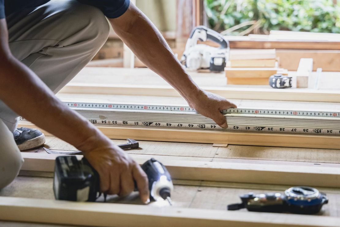 Une personne mesure un tasseau de bois avec un mètre ruban, présence d'outils électriques