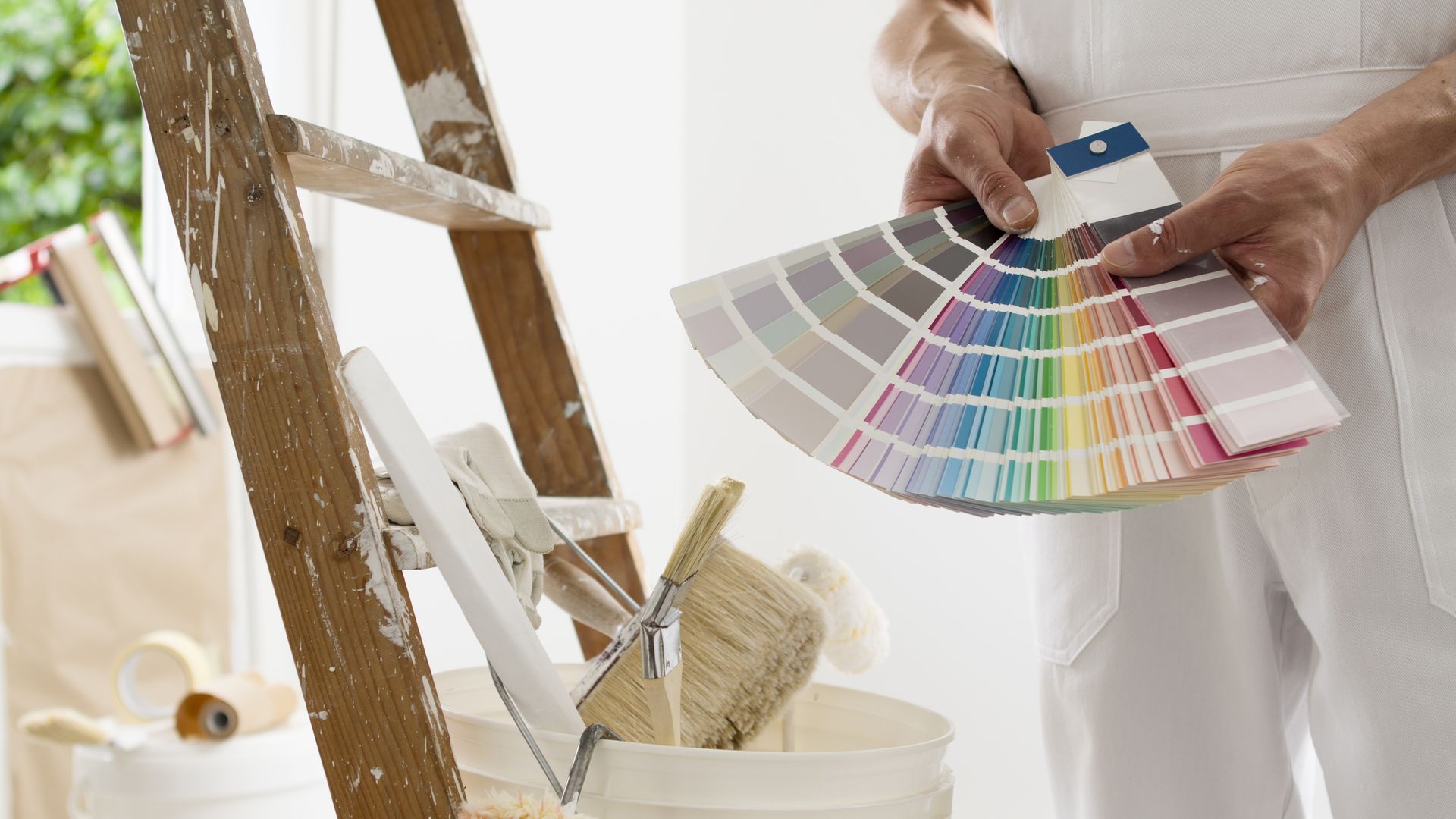 Une personne tenant un éventail d'échantillons de peinture à côté d'une échelle et d'un pot de peinture dans une pièce en cours de peinture
