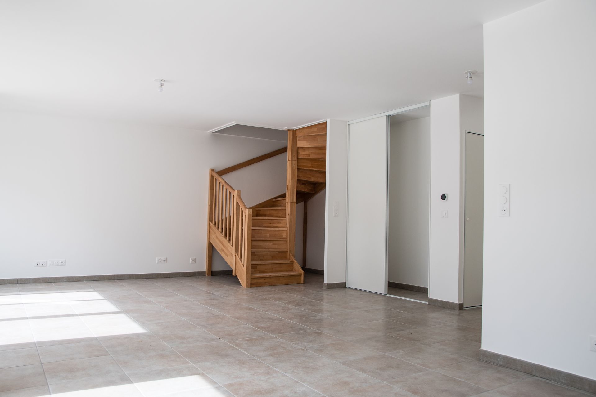 Pièce blanche vide avec sol carrelé, escalier en bois et placards intégrés