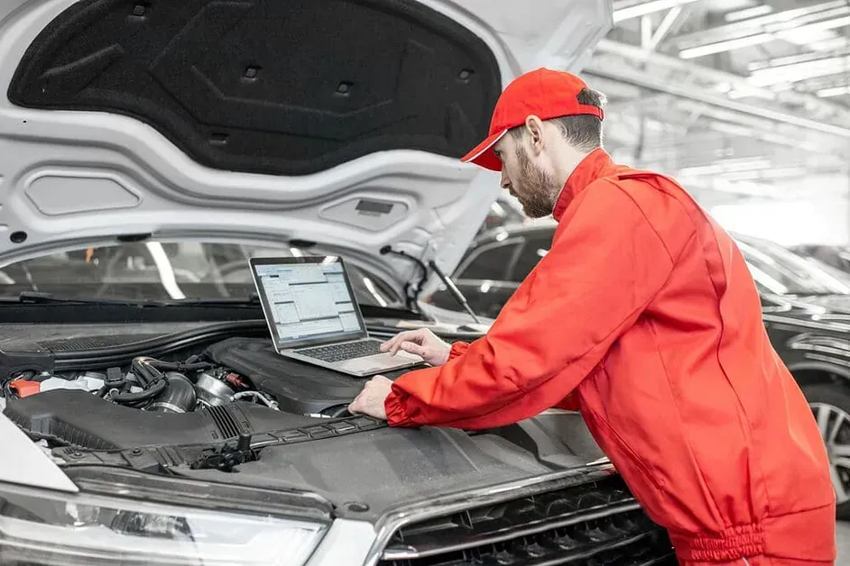 Un hombre está trabajando en el motor de un automóvil mientras usa una computadora portátil.