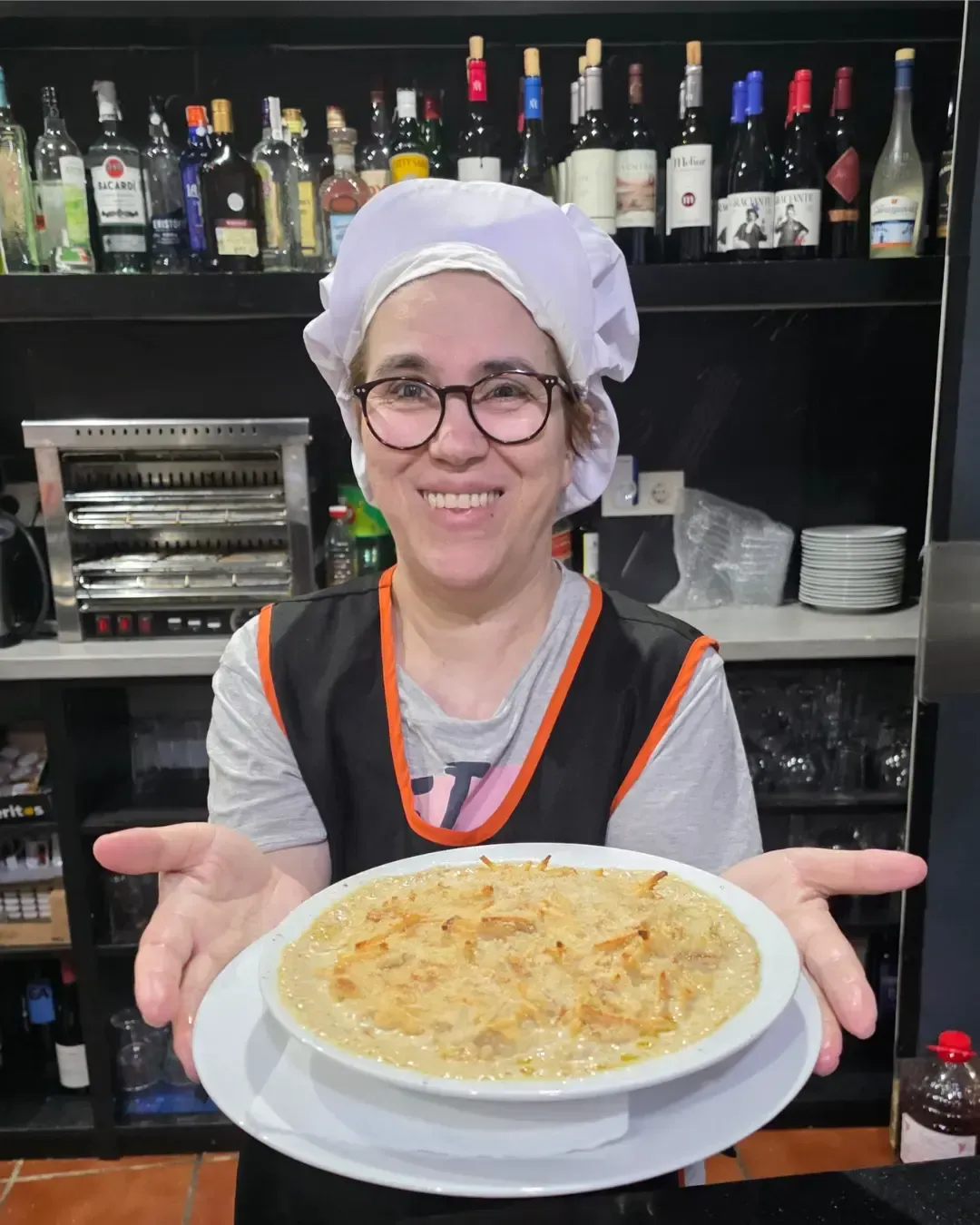 Chef sonriente presentando un tazón de pasta cremosa en una cocina con botellas en estantes detrás de ella.