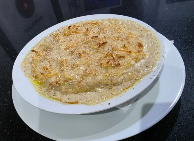 Plato cremoso horneado en un tazón blanco sobre un plato blanco, cubierto con migas doradas sobre una encimera oscura.