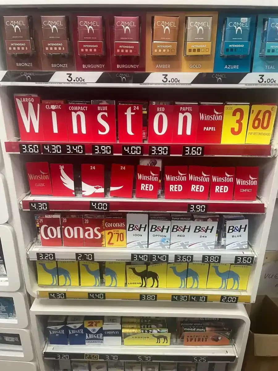 Un expositor en una tienda con estantes de varias marcas de cigarrillos y etiquetas de precio impresas debajo.