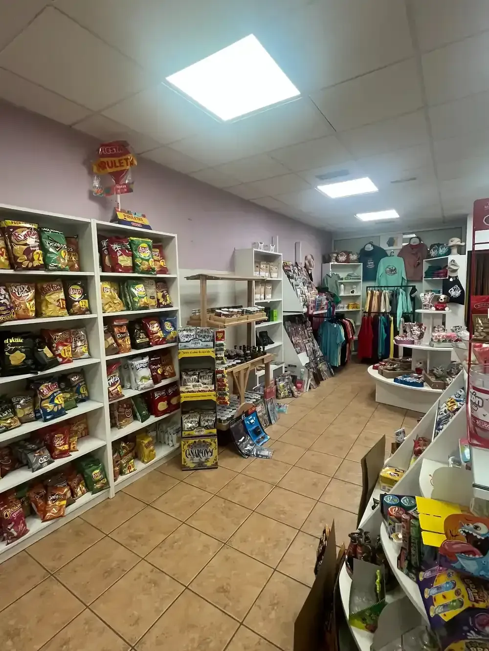 Interior de una tienda con estantes repletos de aperitivos envasados, ropa expuesta y suelo de baldosas.