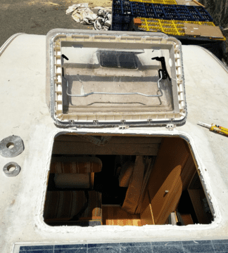 Escotilla de barco abierta sobre una superficie blanca, que deja ver un interior de madera; la escotilla está abierta y colocada encima.