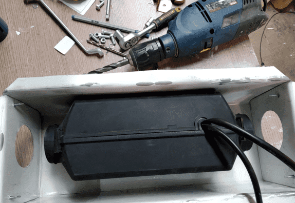 Calentador negro dentro de una caja metálica blanca con un taladro al fondo. Del calentador salen cables.