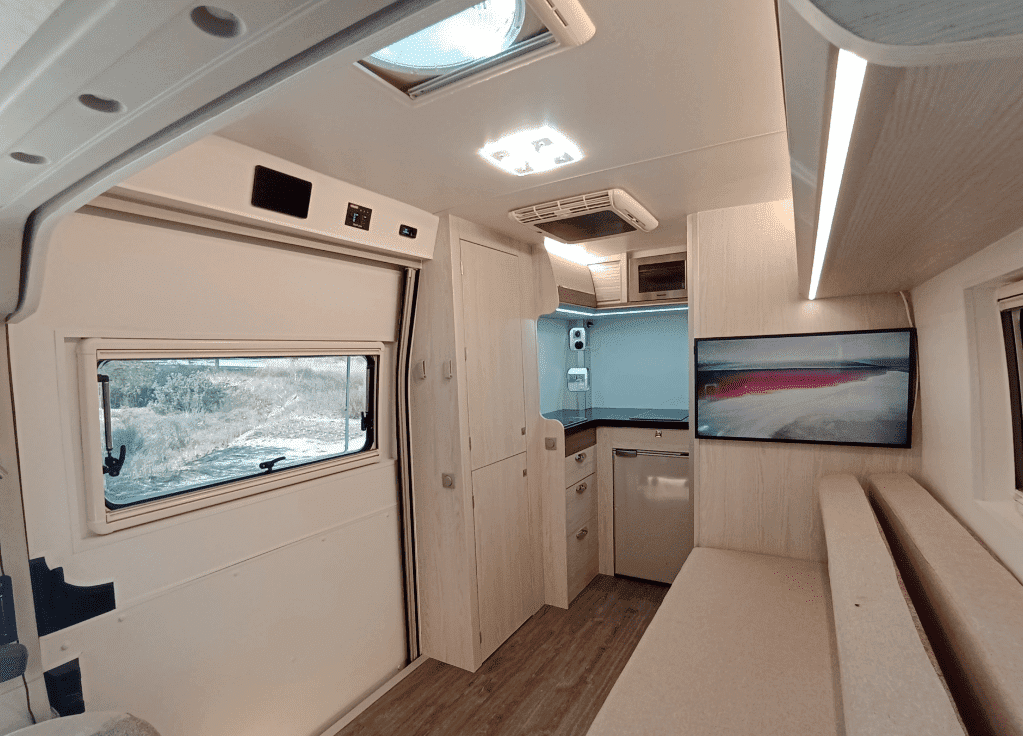 Interior de una moderna furgoneta camper con paredes blancas, armarios empotrados, una pequeña cocina y una ventana con vistas al exterior.
