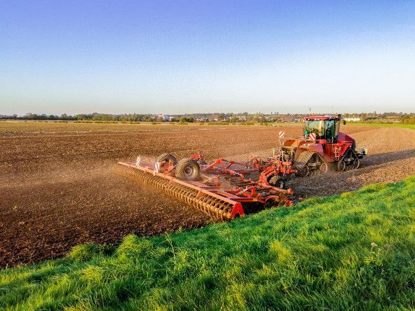 Un tractor ara un gran campo marrón bajo un cielo azul claro, con hierba verde en primer plano.