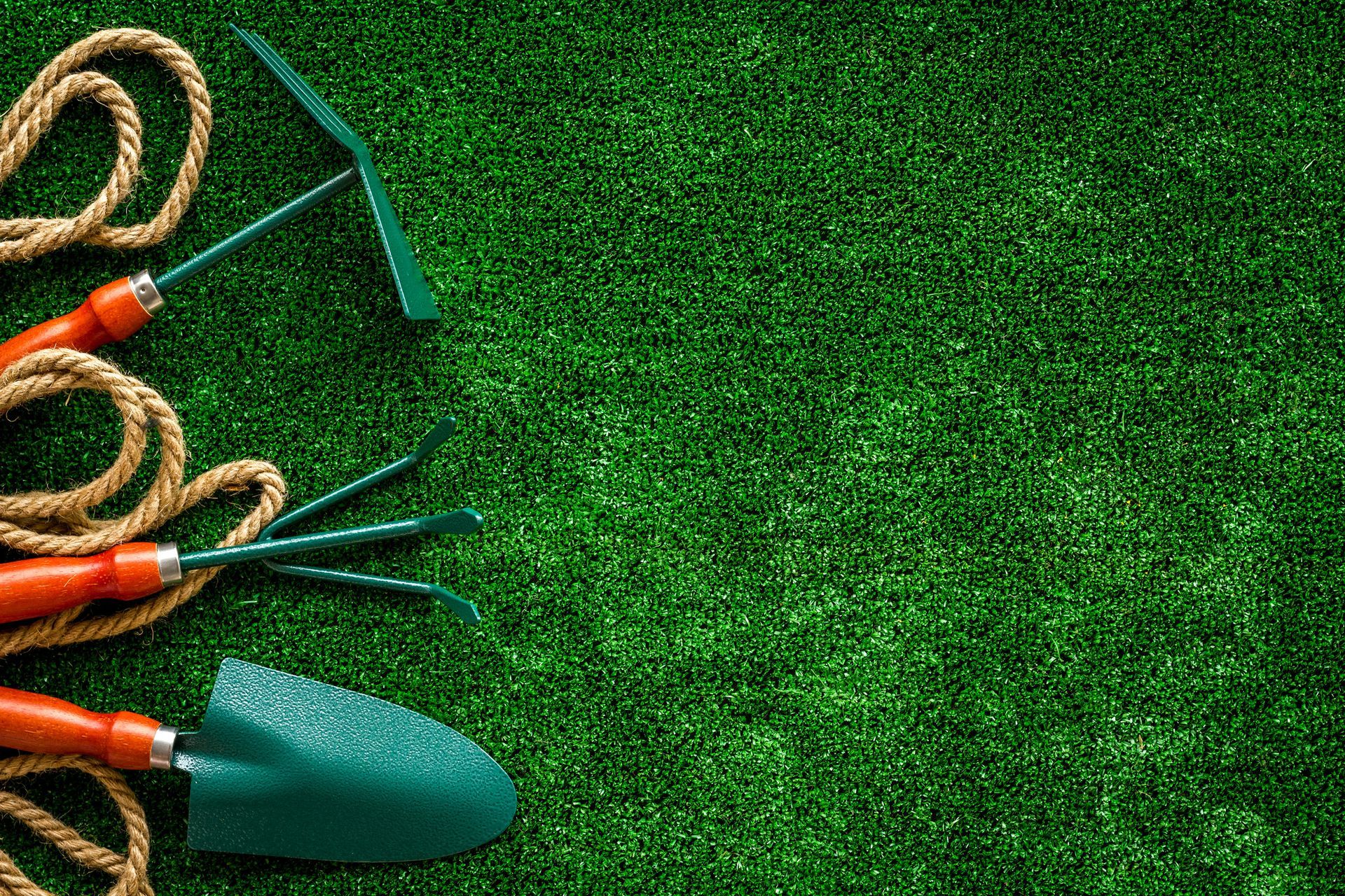 Herramientas de jardinería (paleta, rastrillo, cultivador) y cuerda sobre césped verde.