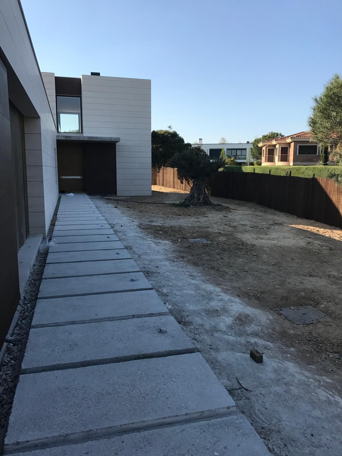Camino de hormigón al lado de una casa blanca moderna, que conduce a un patio con tierra y un seto.