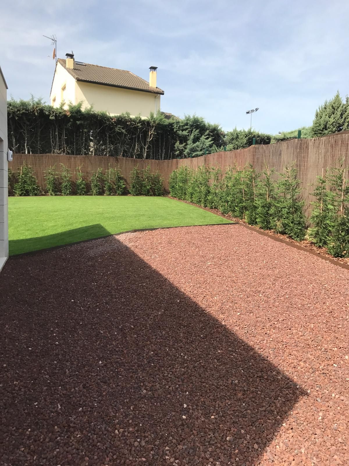 Un patio trasero con grava roja, césped verde y una valla alta de madera. Se ve una casa al fondo.