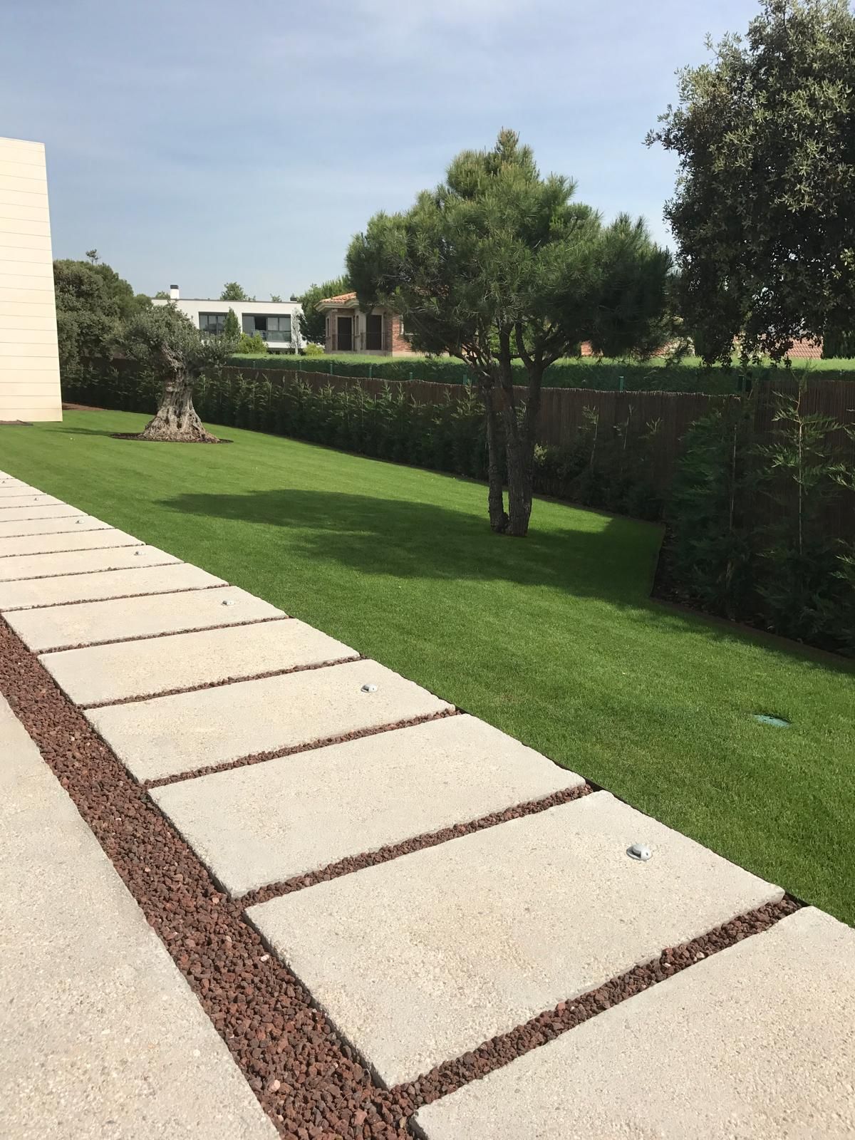 Pasarela de hormigón junto a un césped, árboles y setos contra un cielo brillante.