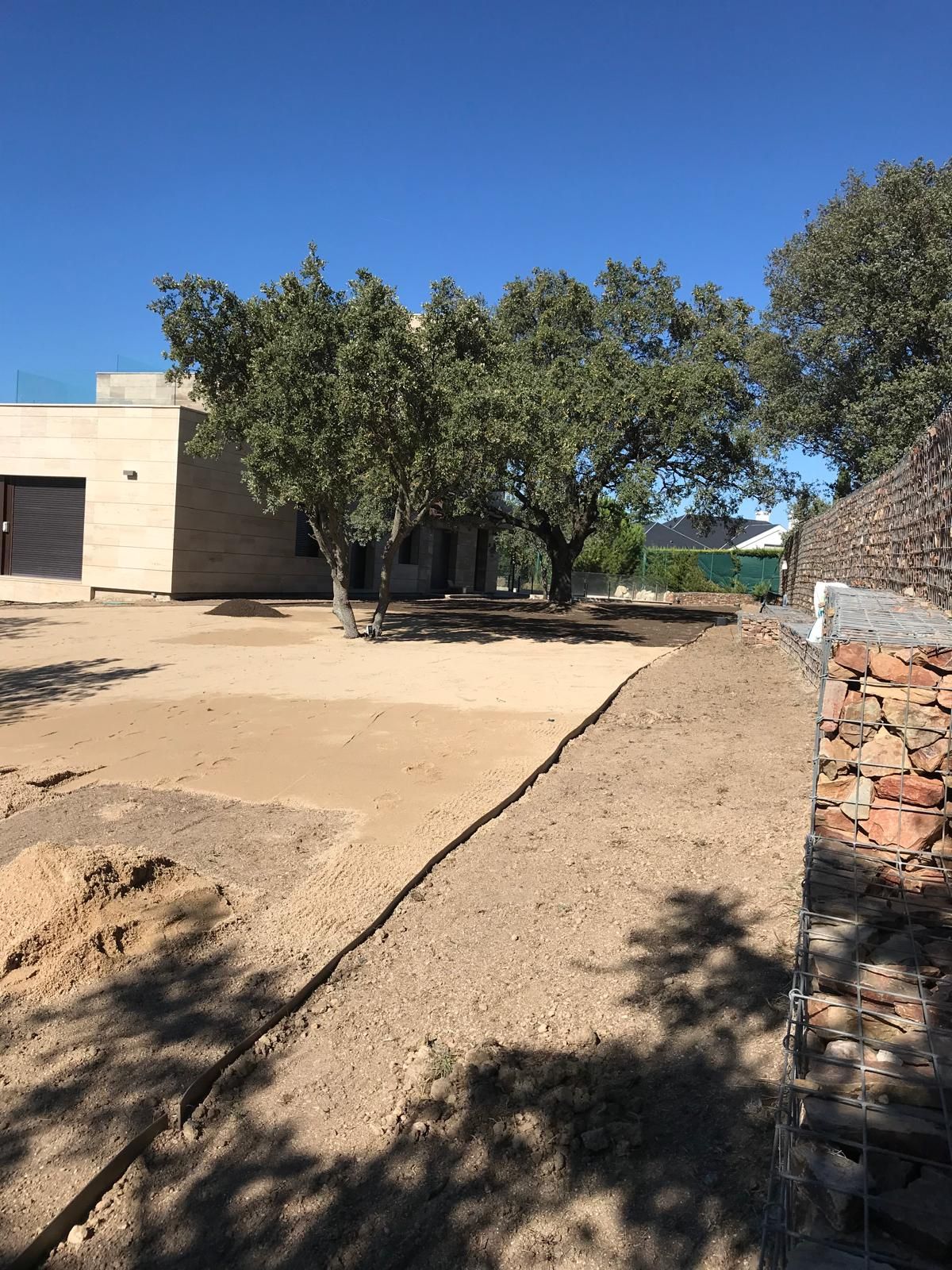 Una escena soleada al aire libre con un edificio beige, árboles y un camino de tierra. Un muro de piedra a la derecha.