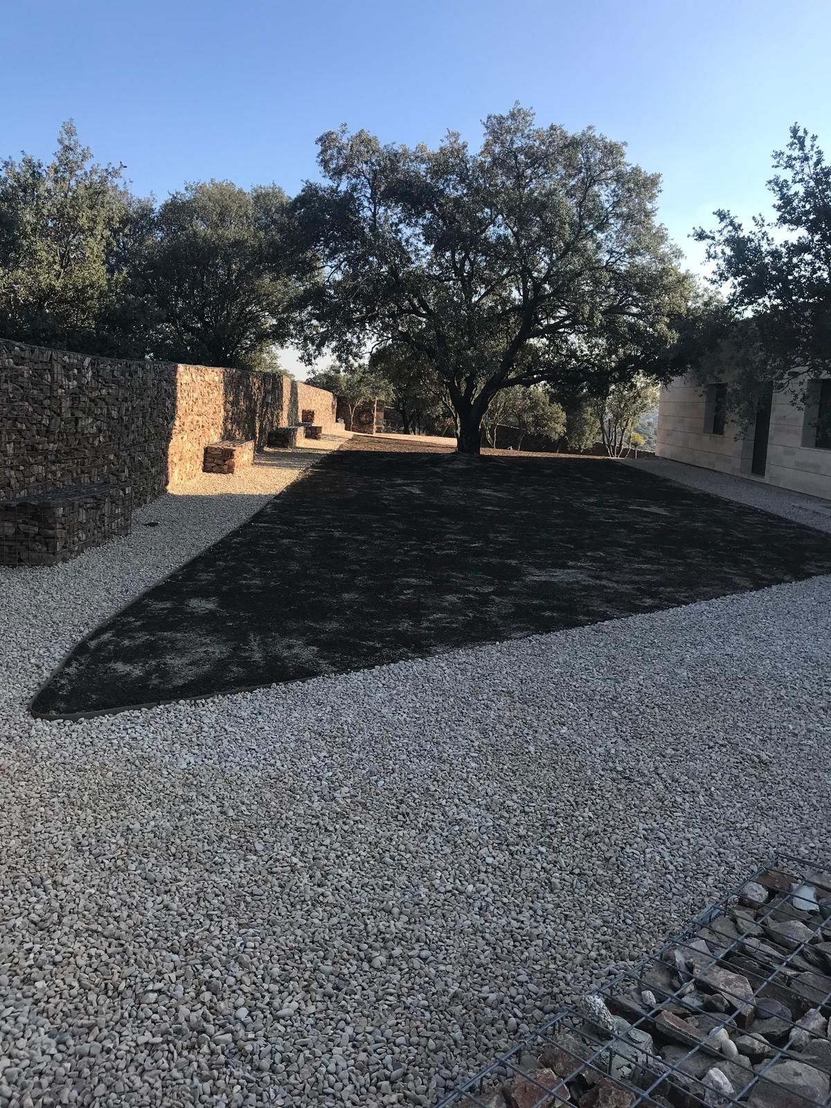 Un camino de grava conduce a una zona cubierta de hierba bajo un gran árbol, bordeada por muros de piedra y un cielo despejado.