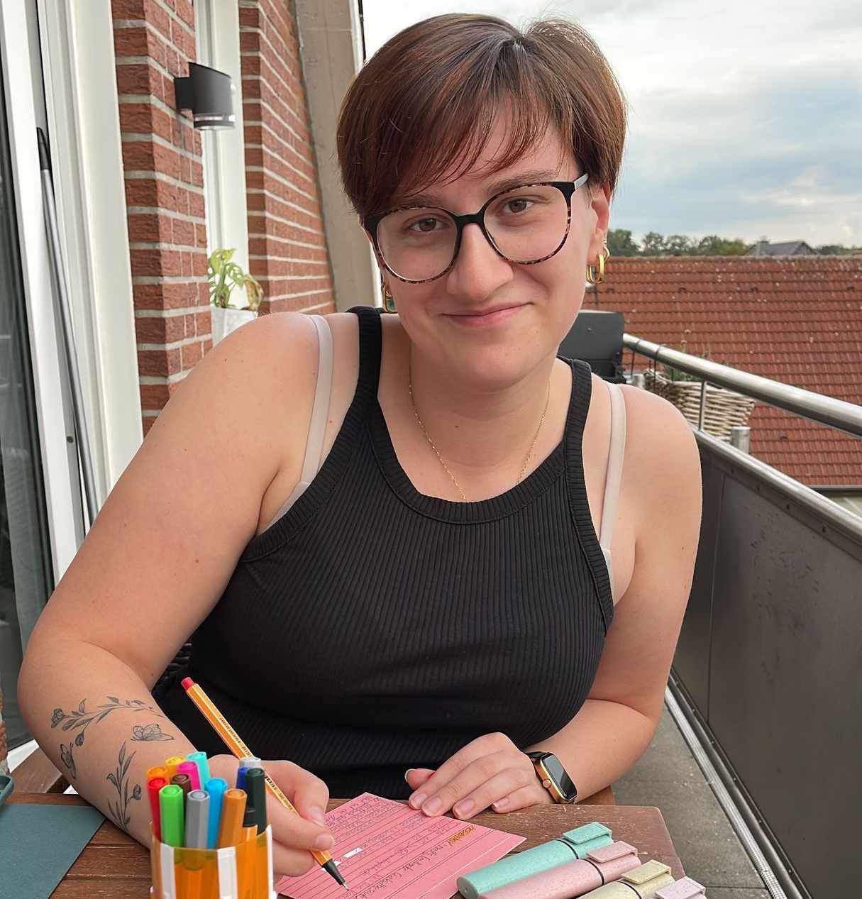 Eine Frau mit Brille und schwarzem Tanktop sitzt an einem Tisch und schreibt auf ein Blatt Papier.