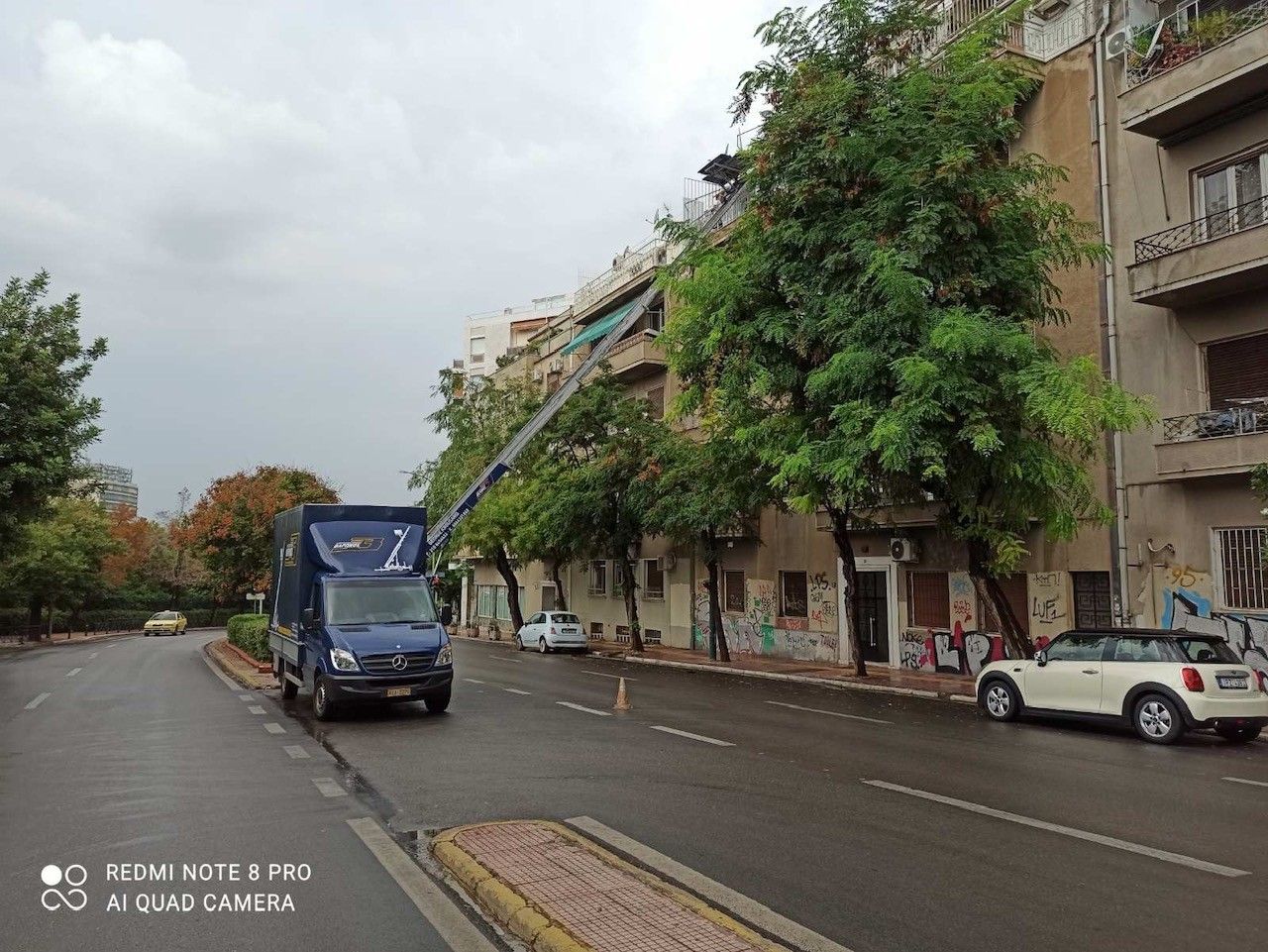 Μπλε φορτηγό πολλαπλών χρήσεων με υπερυψωμένο βραχίονα σε έναν βρεγμένο δρόμο της πόλης δίπλα σε πολυκατοικίες και δέντρα