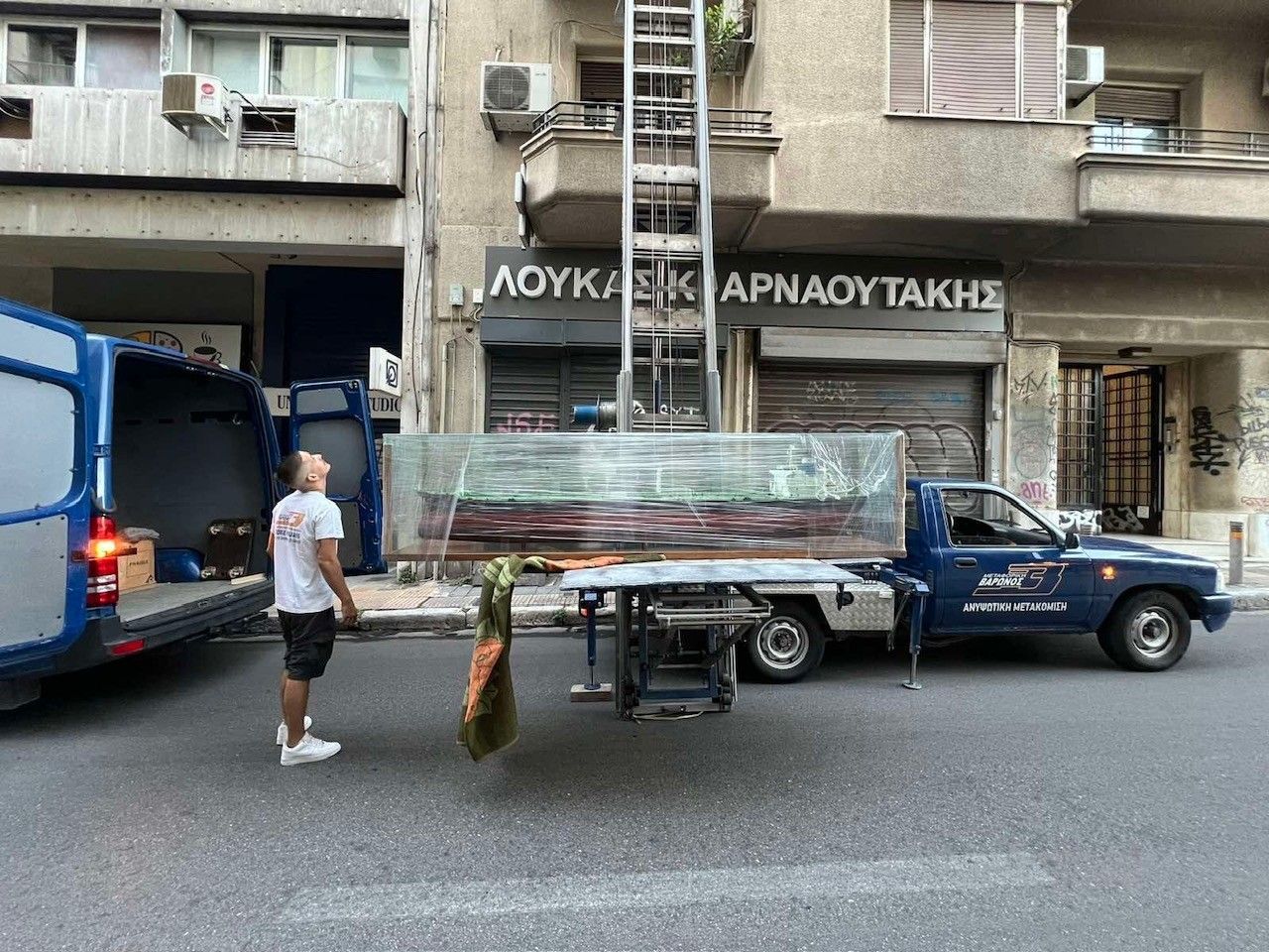 Σκηνή δρόμου με ένα μπλε φορτηγό που μεταφέρει ένα τυλιγμένο φορτίο και έναν άντρα να στέκεται δίπλα του μπροστά σε μια πολυκατοικία