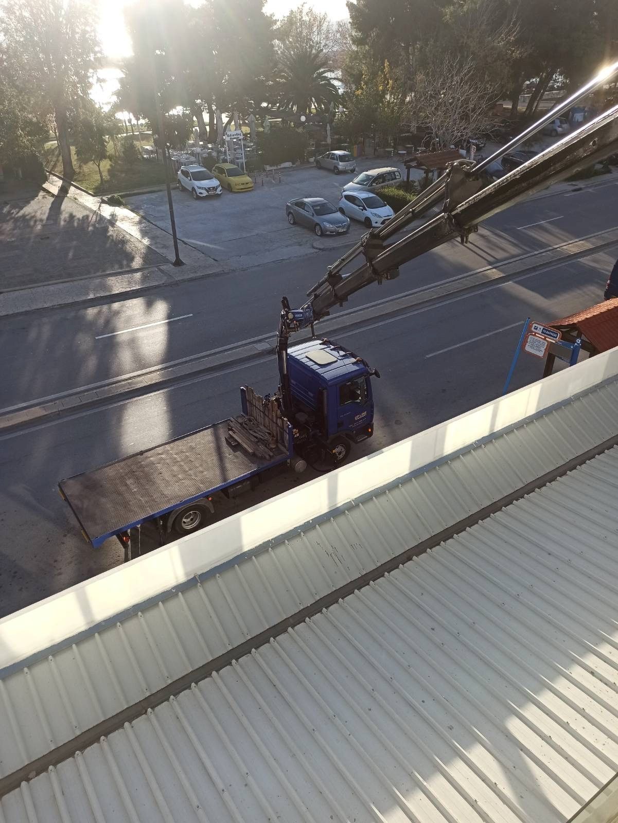 Μπλε φορτηγό με εκτεταμένο γερανό παρκαρισμένο δίπλα σε δρόμο, όπως φαίνεται από ψηλά με δέντρα και αυτοκίνητα κοντά