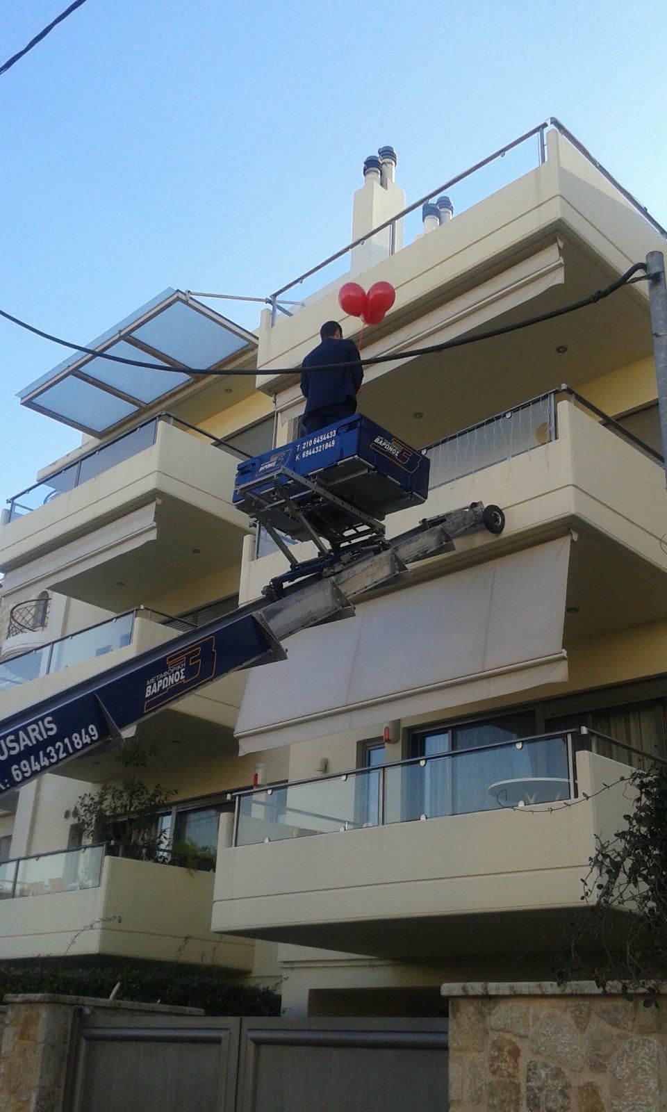 Εργάτης σε μπλε ασανσέρ που επισκευάζει ένα μπαλκόνι σε μια μπεζ πολυκατοικία κάτω από μια κόκκινη διακόσμηση καρδιάς.