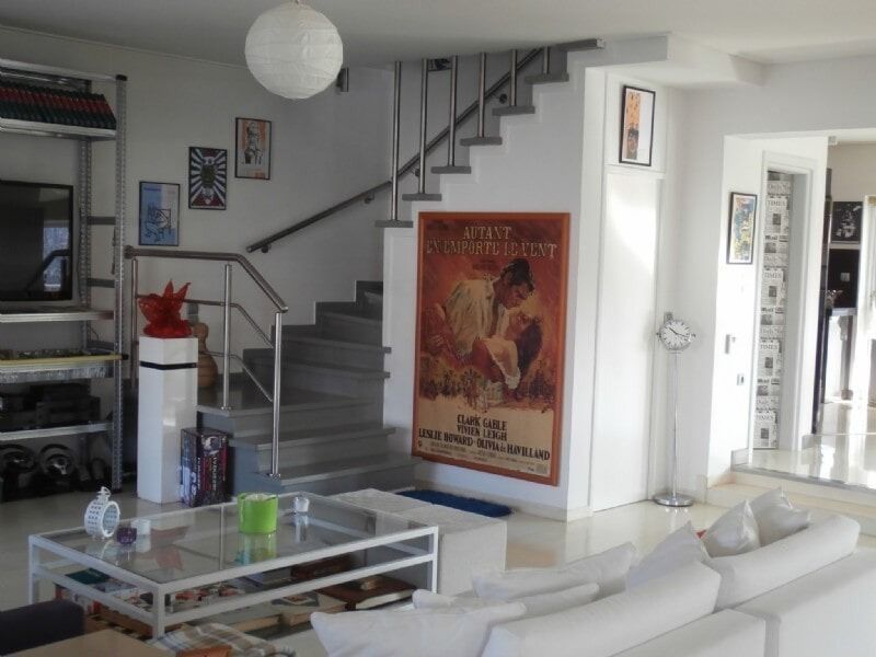 Φωτεινό σαλόνι με λευκό καναπέ, γυάλινο τραπεζάκι σαλονιού, σκάλες και έργα τέχνης στον τοίχο.