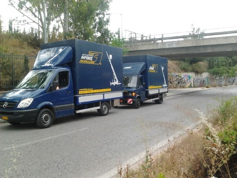 Μπλε φορτηγό διανομής με τρέιλερ οδηγεί σε δρόμο κάτω από μια αερογέφυρα κοντά σε τοίχους καλυμμένους με γκράφιτι.