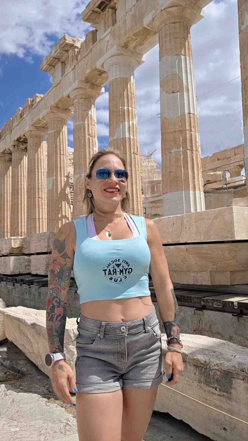 Mujer con gafas de sol y top corto azul de pie frente a antiguas columnas de piedra bajo un cielo brillante.