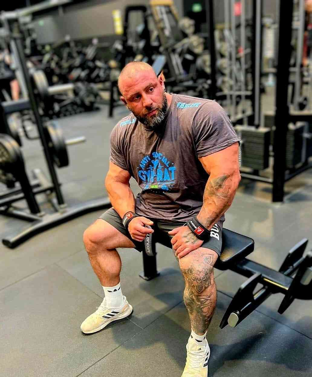 Hombre sentado en un banco de gimnasio, con camiseta gris y pantalones cortos, con máquinas de pesas al fondo.