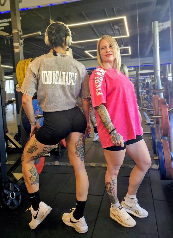 Dos personas posando en un gimnasio, una vestida de gris y negro, la otra de rosa, ambas con auriculares y zapatillas deportivas.