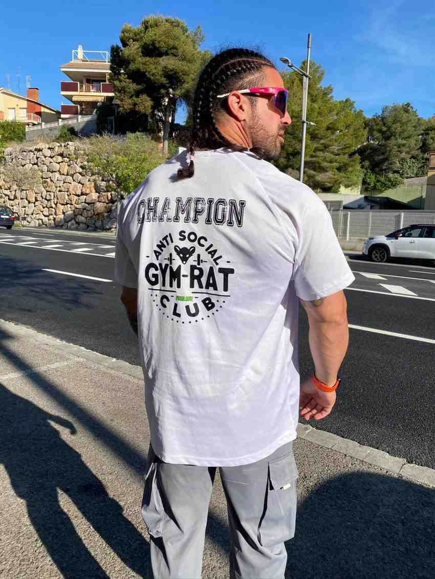 Hombre de pie al aire libre, con camisa gris y pantalones cargo, y gafas de sol, cerca de un muro de piedra.