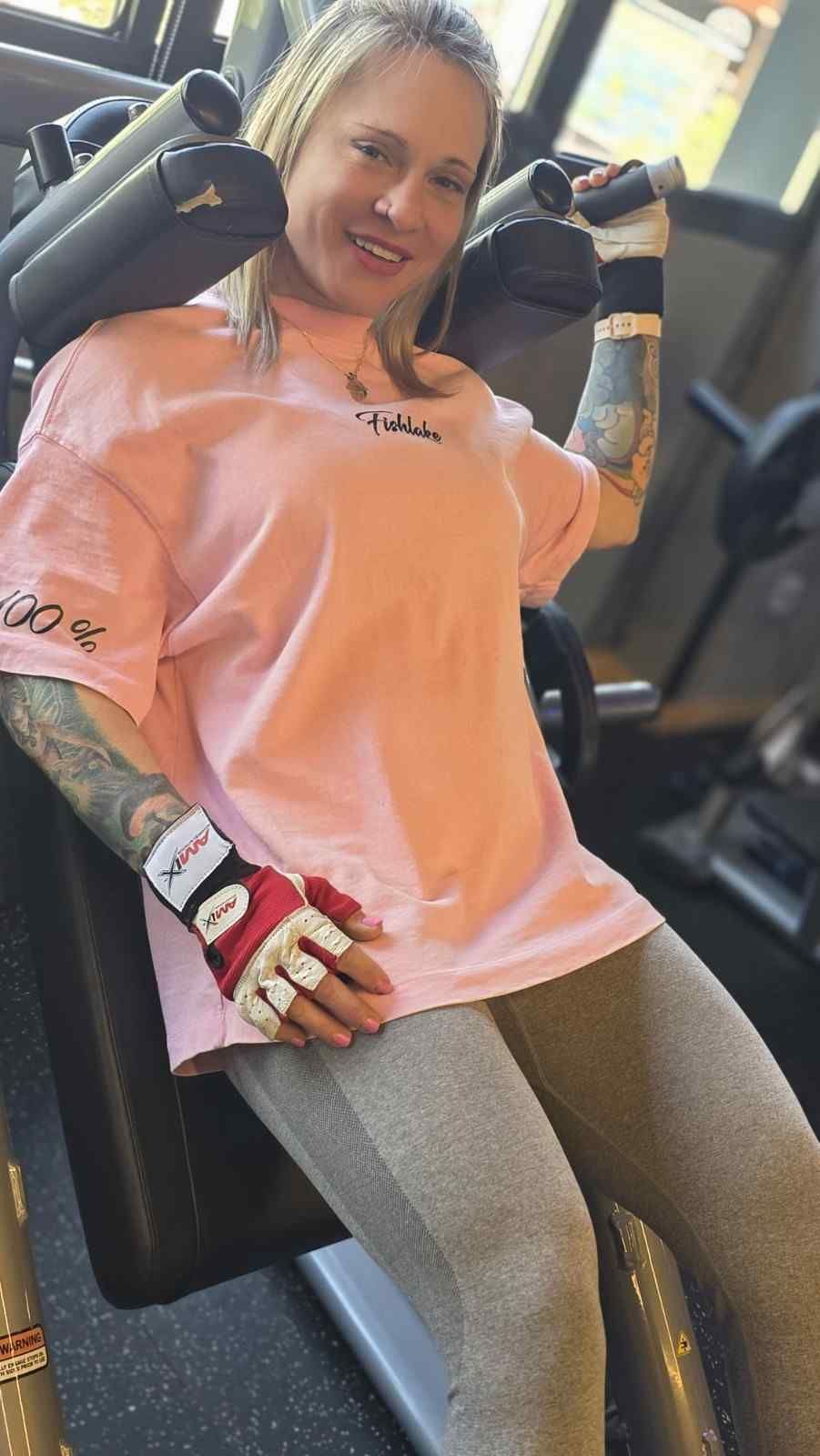 Mujer con camisa rosa y mallas grises sonriendo en una máquina de ejercicios del gimnasio, con guantes de boxeo.