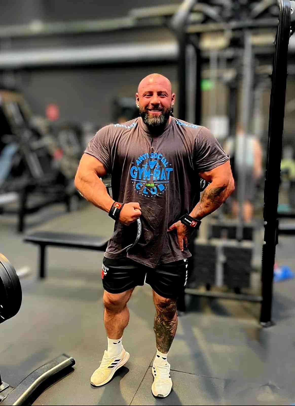 Hombre sentado en un banco de gimnasio, con camiseta gris y pantalones cortos, con máquinas de pesas al fondo.