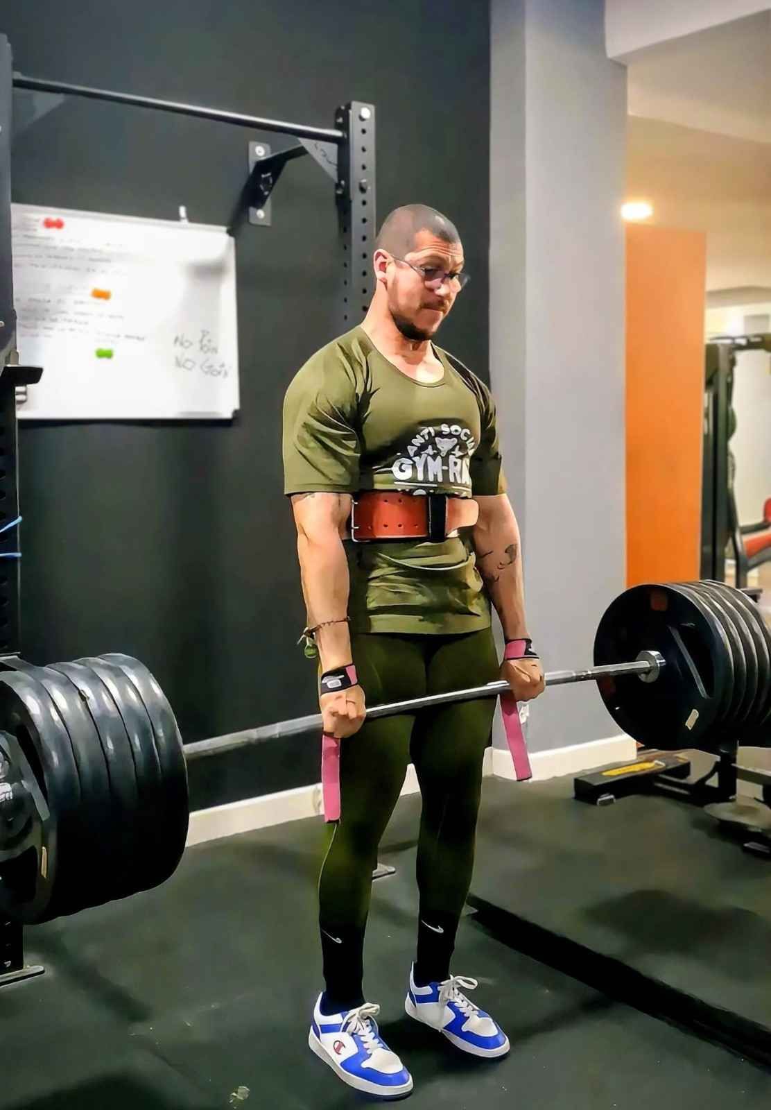Persona levantando una barra pesada en un gimnasio, vestida con ropa deportiva verde y correas de levantamiento.