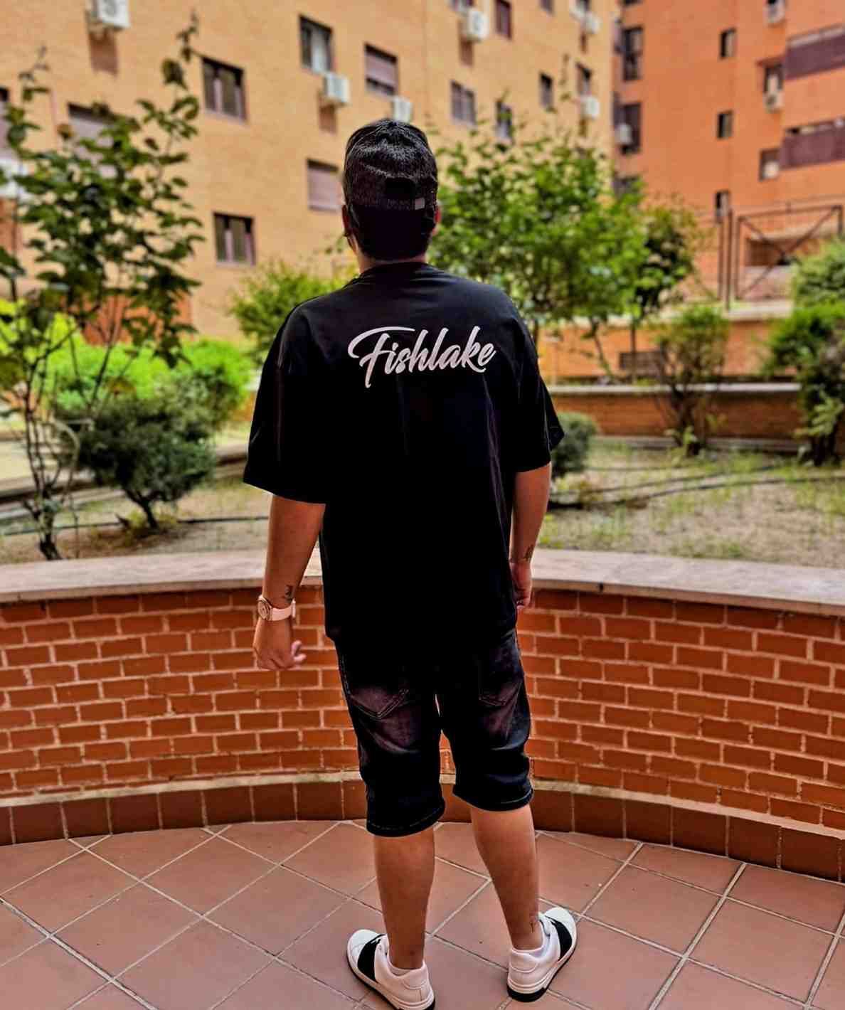 Persona vestida de negro de pie en un patio, vista desde atrás, con edificios de apartamentos y vegetación al fondo.
