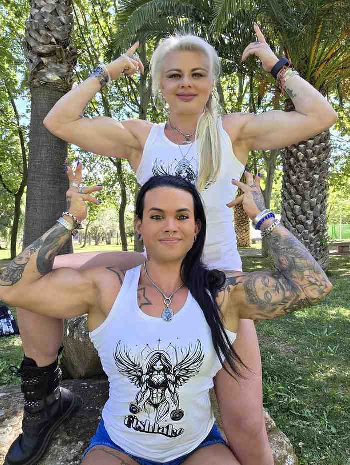 Dos mujeres tatuadas posando al aire libre, flexionando los brazos con blusas blancas y pantalones negros.