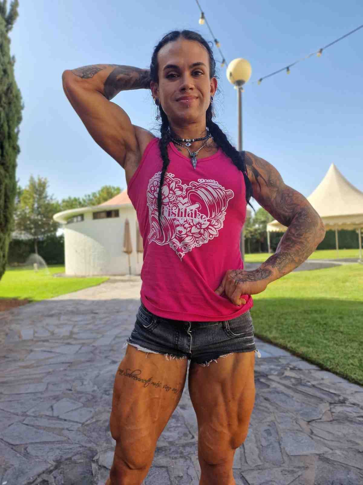 Persona atlética posando al aire libre con una camiseta rosa y pantalones cortos negros, con el pelo trenzado y brazos.