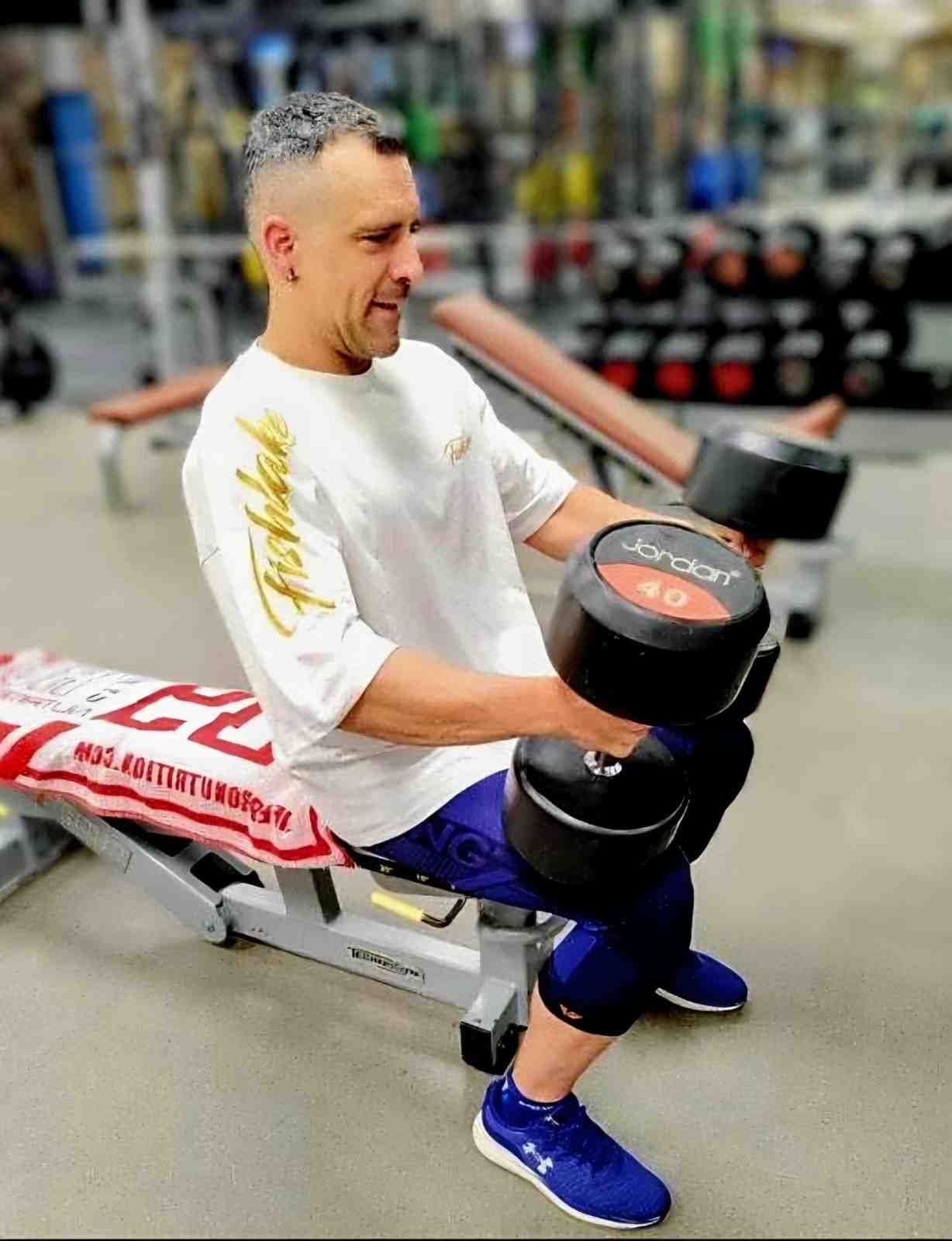 Hombre levantando una mancuerna sentado en un banco de gimnasio, vistiendo una camisa blanca y pantalones cortos azules.