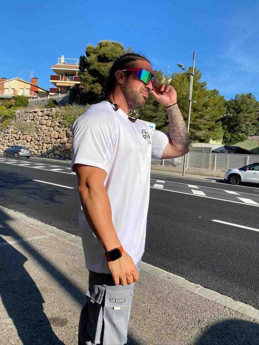 Un hombre con camiseta blanca y pantalones cortos grises está de pie al borde de una carretera soleada.