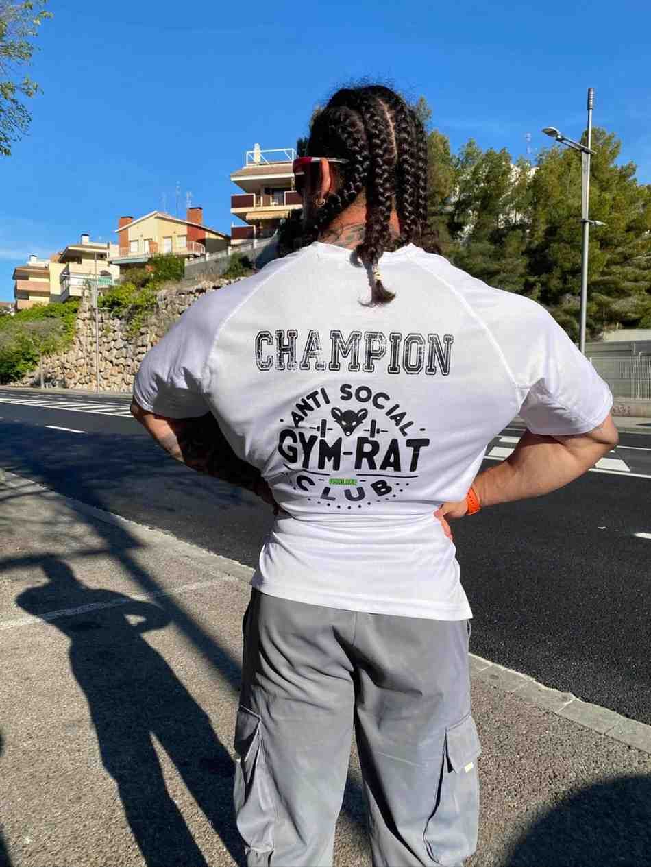 Persona de pie al borde de la carretera, vista de espaldas, con camiseta blanca y pantalones grises y el pelo trenzado.