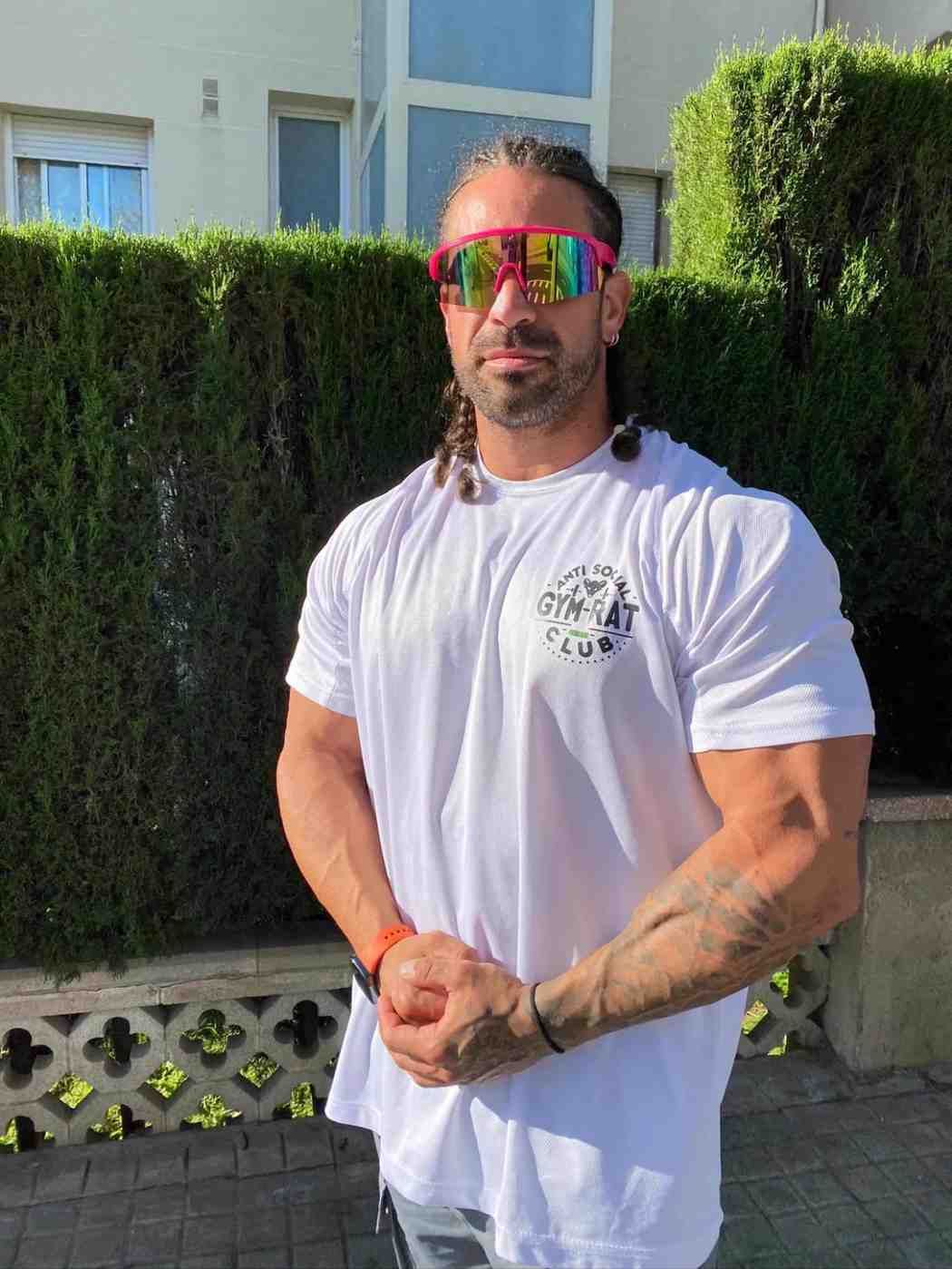 Un hombre está de pie al aire libre, con una camiseta blanca y gafas de sol reflectantes, con los brazos cruzados .