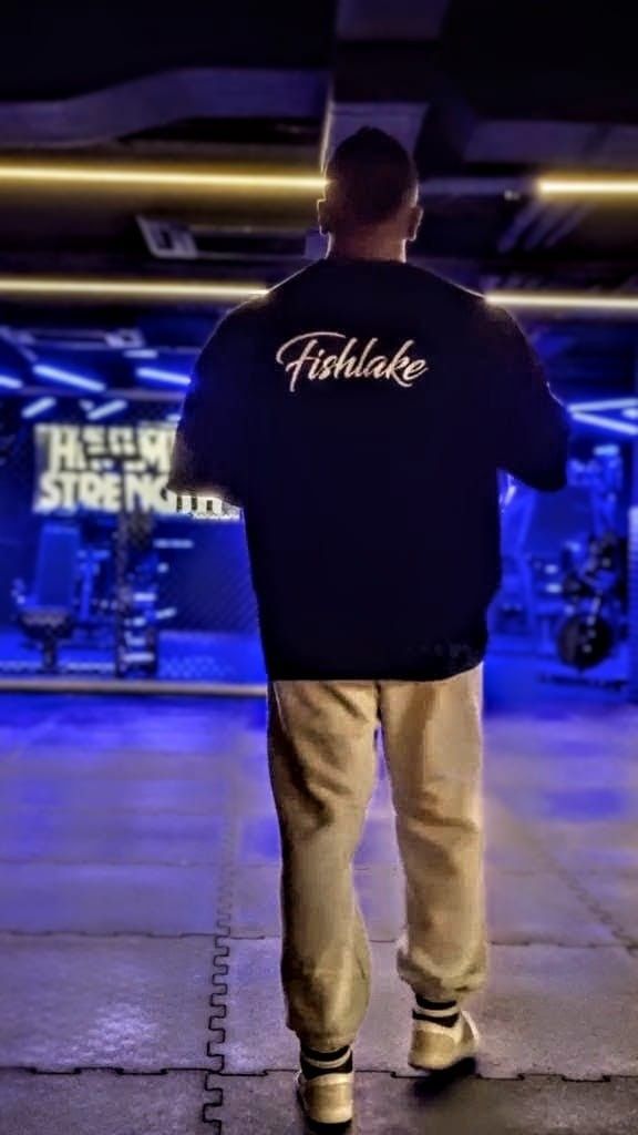 Hombre con chaqueta negra y pantalones color canela de pie en un gimnasio iluminado con luces de neón, visto desde atrás.