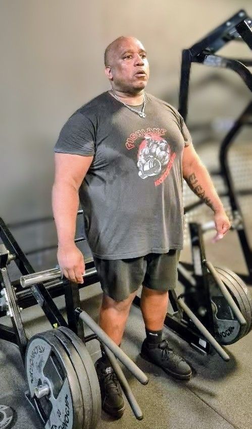 Hombre de pie en un gimnasio junto a una máquina de pesas cargada, vistiendo una camiseta gris y pantalones cortos negros.