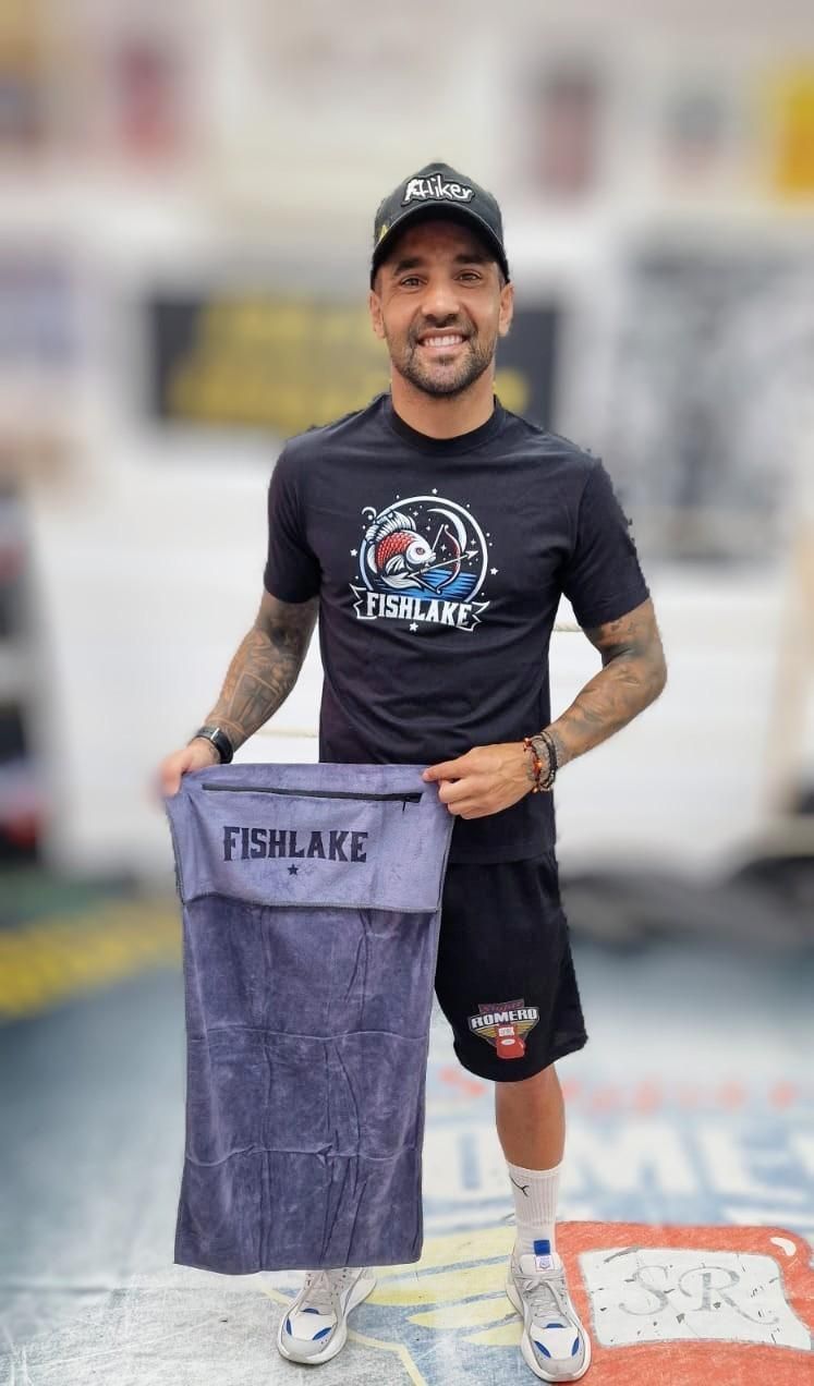 Hombre de pie en un gimnasio, sosteniendo una toalla, vestido con ropa deportiva negra y una gorra, sonriendo.