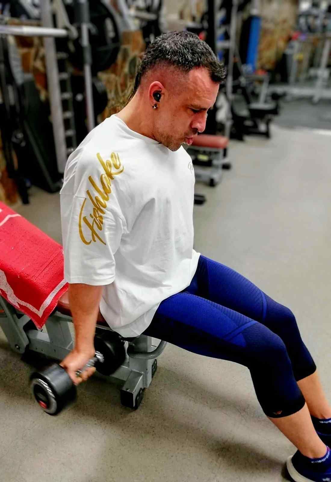 Persona realizando un ejercicio con mancuernas sentada en un gimnasio, vestida con una camiseta blanca y mallas azules.