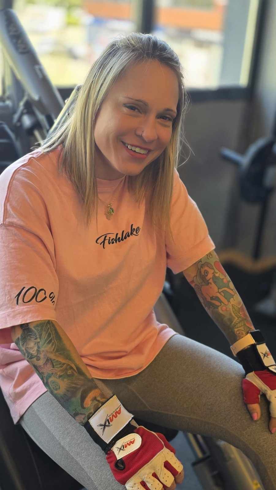 Una persona sonriente con una camisa rosa y guantes de boxeo rojos sentada en una bicicleta estática en el interior.
